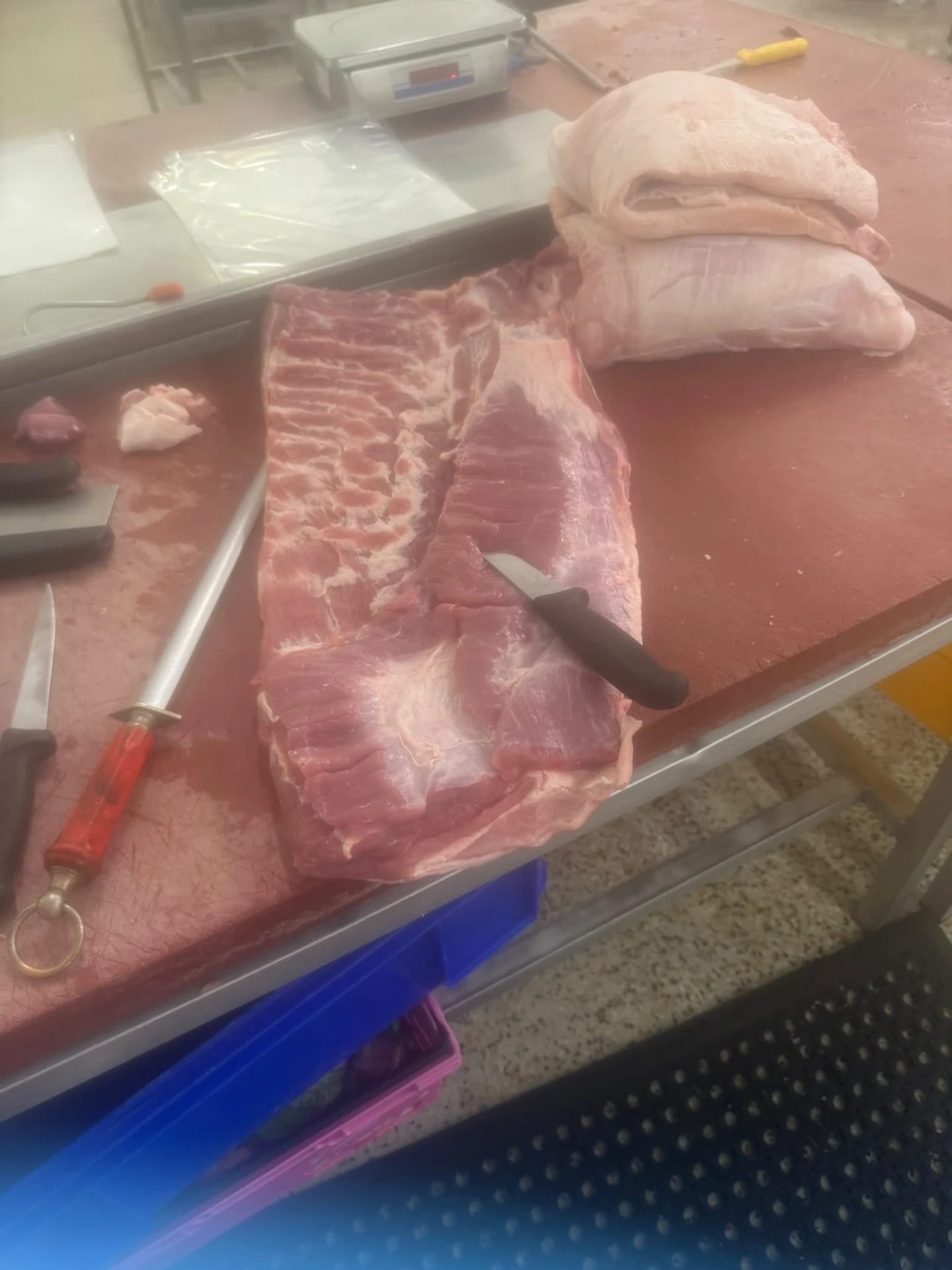 A pork loin being prepared by hand on the butcher's block, with the butcher's knives and steel alongside
